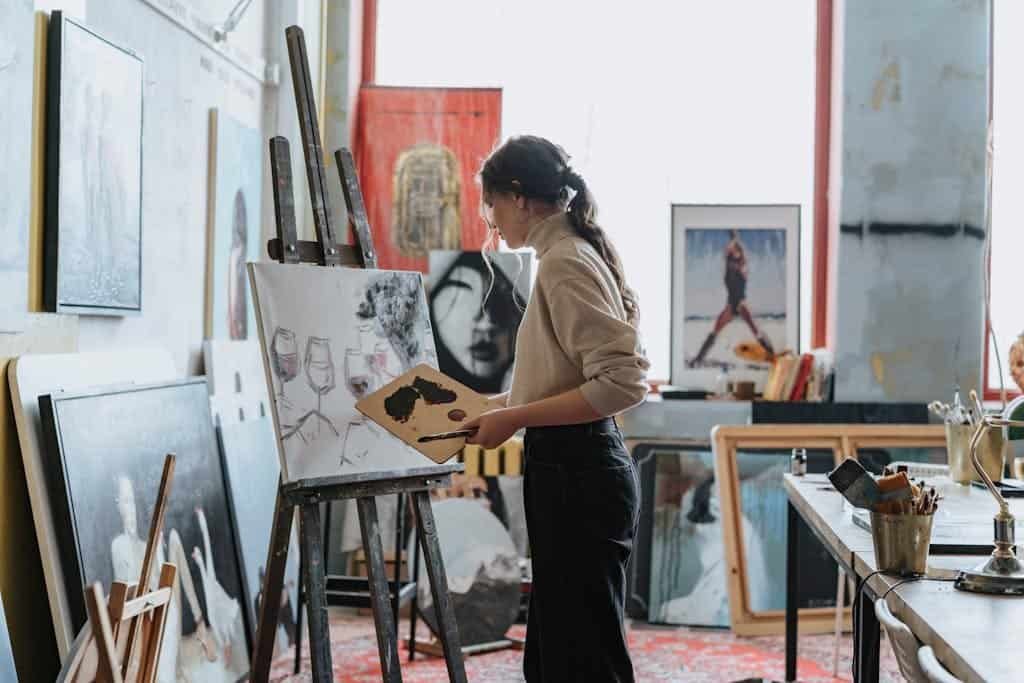 A woman artist works on a painting in a creative studio space filled with art and light.